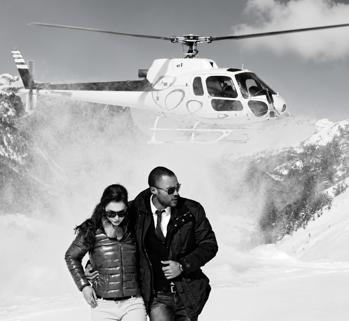 A black and white lifestyle photograph of a couple walking in the snow with a helicopter flying overhead in the background.